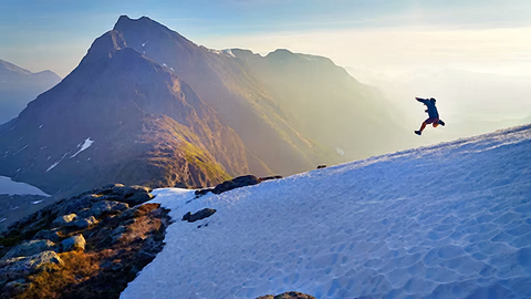 A lone figure leaping across a sunlit, snow-covered mountain ridge