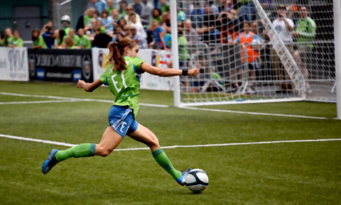 A soccer player striking the ball in front of a crowded stadium goal
