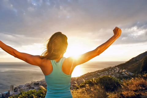 A person standing on a hillside at sunrise with both arms raised, looking out over a coastal city