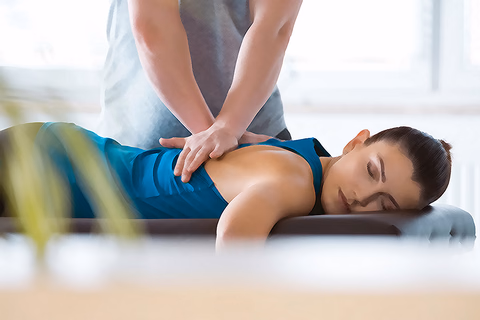 A practitioner treats the mid-back of a patient lying face down in a bright, sunlit treatment room